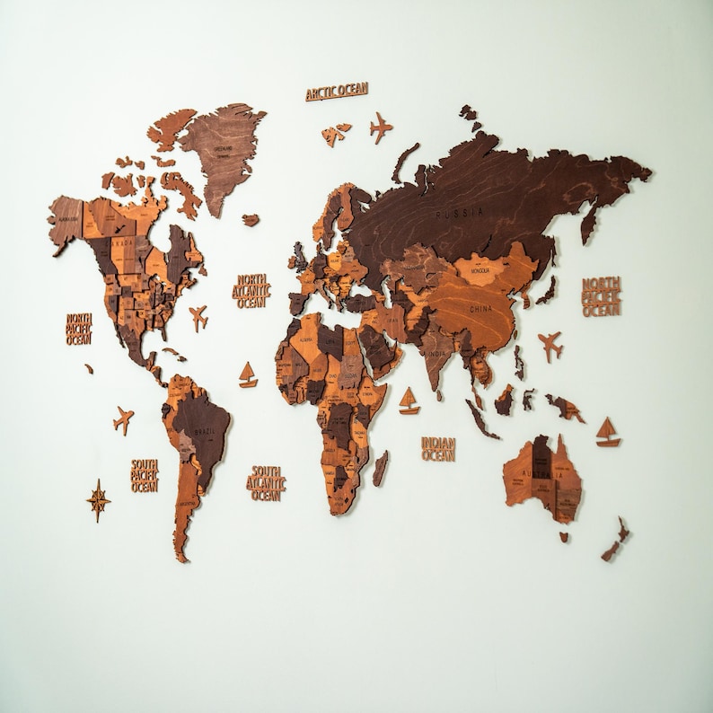 Large 3D wooden world map wall decor in mixed brown tones — laser-cut birch plywood with country names, ocean labels, and decorative wooden planes and sailboats