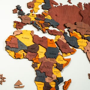Detail shot of colorful 3D wooden world map — laser-engraved country names across Europe, Africa and Middle East in vibrant yellow, orange, brown and grey wood tones with red location pins