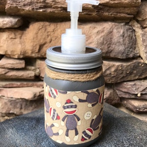 May include: Gray glass soap dispenser with a silver lid and a pump. The dispenser is decorated with a brown and beige pattern of monkeys wearing red hats.