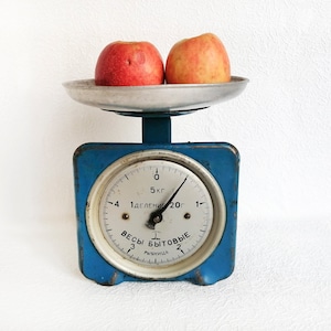 May include: A blue vintage kitchen scale with a white face and black markings. The scale has a silver tray with two red apples on top. The face of the scale reads "Весы бытовые" and "1 деление 20г".