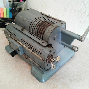 Puede incluir: Calculadora mecánica vintage en tonos grises y azules. El dispositivo presenta múltiples diales numéricos, una manivela y varias palancas. La calculadora tiene una base rectangular y una sección superior cilíndrica, mostrando signos de edad y uso.