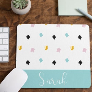 Può includere: Un tappetino per mouse bianco con un bordo inferiore blu chiaro. La parte superiore del tappetino per mouse presenta un motivo ripetitivo di quadrati neri, pennellate rosa e gialle. Il bordo inferiore riporta il nome "Sarah" scritto in corsivo bianco.