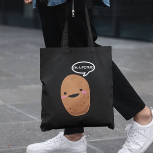 Könnte beinhalten: Eine schwarze Tragetasche mit einer weißen Cartoon-Kartoffel und dem Text "Ich bin eine Kartoffel" in einer Sprechblase. Die Tasche wird von einer Person getragen, die schwarze Hosen und ein blaues Jeanshemd trägt.