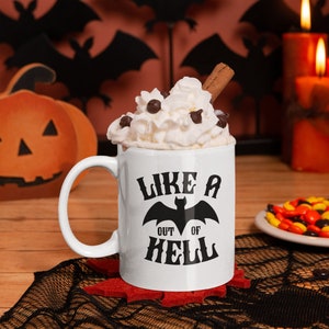 Op de afbeelding: Een witte keramische koffiemok met een zwarte vleermuisafbeelding en de tekst "Like a Out of Hell". De mok is gevuld met slagroom en een kaneelstokje. De mok staat op een houten tafel met een zwarte kantonderzetter en een bord met snoep.