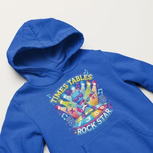 Op de afbeelding: Een koningsblauwe hoodie met een kleurrijke grafische print. Het ontwerp bevat de tekst "TIMES TABLES ROCK STAR" en een stripfiguur die gitaar speelt. De afbeelding bevat muzieknoten, spotlights en wiskundige symbolen.