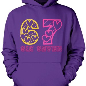 May include: Purple hoodie with a front graphic. The graphic features the number "67" with heart shapes and the words "SIX SEVEN" in pink and yellow. The hoodie has a hood and a front pocket.