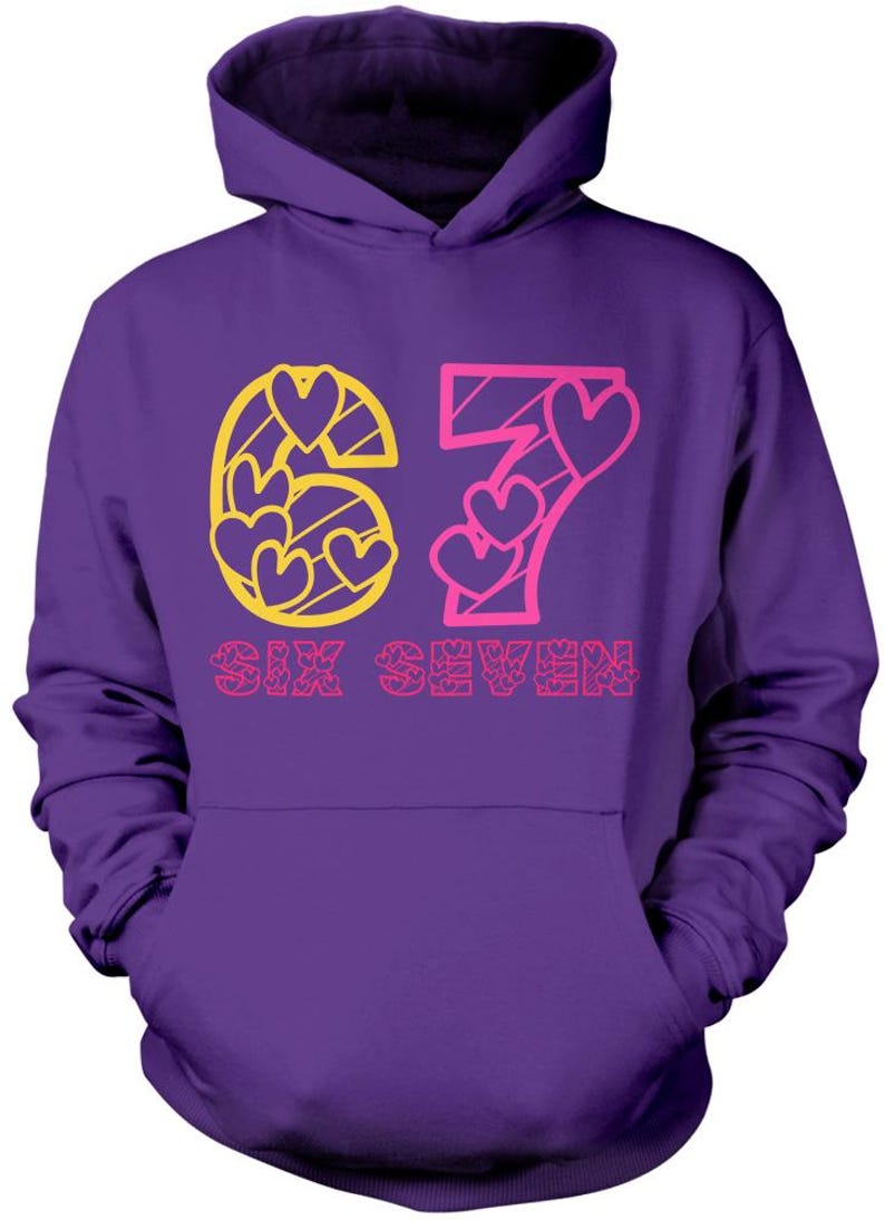 May include: Purple hoodie with a front graphic. The graphic features the number "67" with heart shapes and the words "SIX SEVEN" in pink and yellow. The hoodie has a hood and a front pocket.