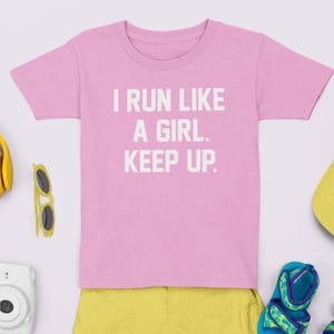 Puede incluir: Camiseta rosa con el texto blanco "I RUN LIKE A GIRL. KEEP UP.". Alrededor, pantalones cortos amarillos, gafas de sol, una cámara, un sombrero y sandalias.