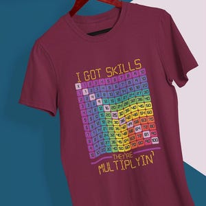 Op de afbeelding: Bordeauxrood t-shirt met de tekst "I GOT SKILLS THEY'RE MULTIPLYIN'" en een kleurrijke vermenigvuldigingstabel. Het t-shirt wordt gepresenteerd op een houten hanger.
