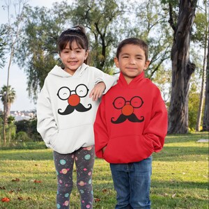Può includere: Due bambini con felpe con cappuccio abbinate con un design stravagante. Una felpa è bianca, l'altra rossa. Ognuna presenta un volto di cartone animato con occhiali rotondi, un naso rosso e baffi neri. I bambini sono in un'area erbosa.