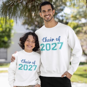 Könnte beinhalten: Zwei weiße langärmelige Sweatshirts mit Rundhalsausschnitt, die den Text "Choir of" in blauer Schreibschrift über der Jahreszahl "2027" in großen, türkisfarbenen Blockbuchstaben mit dünner Umrandung zeigen. Die Kleidungsstücke haben ein schlichtes Design.