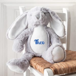 Op de afbeelding: Een grijze pluche bunny met witte oren en een witte vlek op zijn borst. De bunny zit op een witte stoel met een gevlochten zitting. De naam "Theo" is in blauw geborduurd op de witte vlek.