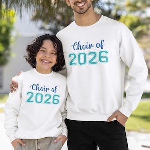 Op de afbeelding: Twee mensen dragen witte sweatshirts met de tekst "Choir of 2026" in blauw script en turquoise blokletters. De sweatshirts hebben een ronde hals. Eén persoon heeft zijn arm om de schouder van de ander.