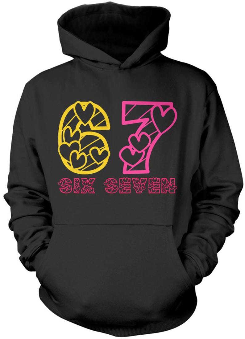 May include: Black hooded sweatshirt with the number "67" in yellow and pink. The "6" is filled with yellow hearts, and the "7" has pink hearts. The words "SIX SEVEN" are printed in pink below the numbers.