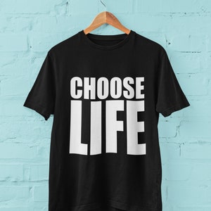 May include: Black t-shirt with white text that reads "CHOOSE LIFE" hanging on a wooden hanger against a light blue brick wall.