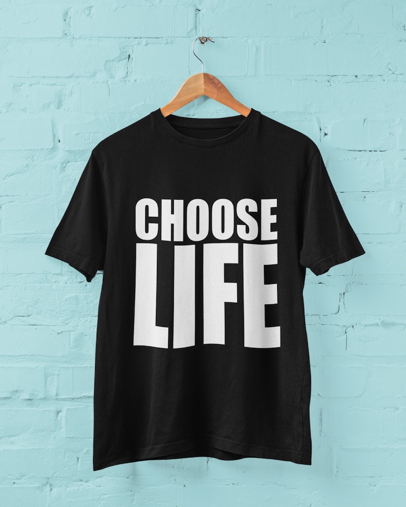 May include: Black t-shirt with white text that reads "CHOOSE LIFE" hanging on a wooden hanger against a light blue brick wall.
