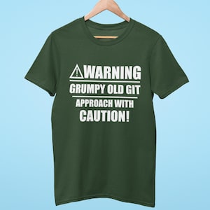 Può includere: Una maglietta verde scuro con la scritta bianca "WARNING GRUMPY OLD GIT APPROACH WITH CAUTION!". La maglietta è appesa a una gruccia di legno su uno sfondo azzurro chiaro. Il testo è in grassetto e senza grazie.