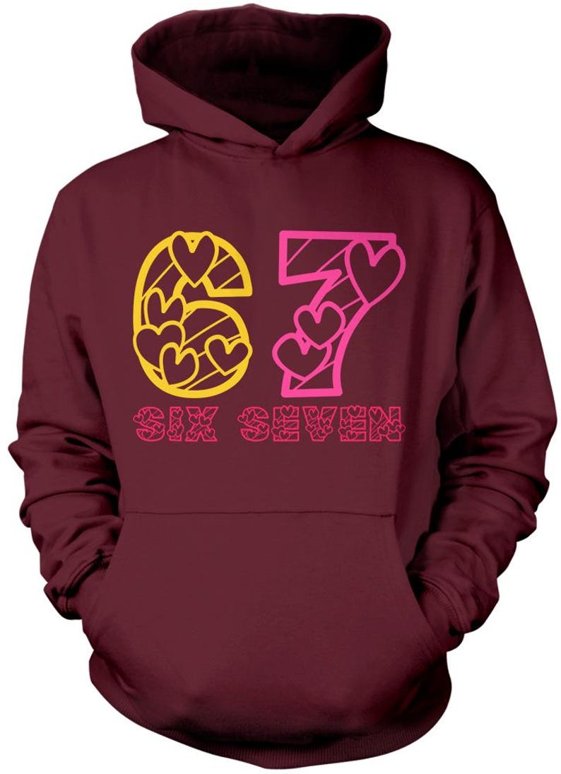 May include: Burgundy hoodie with a front graphic. The graphic features the number 67 in yellow and pink, with heart shapes inside the numbers. The words "SIX SEVEN" are below the numbers in pink.