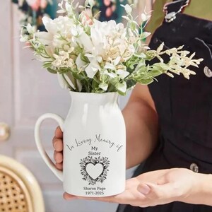 Op de afbeelding: Een witte keramische vaas met een handvat, gevuld met witte en groene bloemen. De vaas heeft een hartvormig bloemmotief en de tekst "In Loving Memory of My Sister Sharon Page 1971-2025".