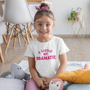 Puede incluir: Una camiseta blanca con el texto "A Little Bit Dramatic" en rosa. La camiseta la lleva una niña con un accesorio para el pelo rosa. La niña sonríe y hay peluches en primer plano.