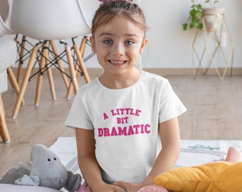 May include: A white t-shirt with the text "A Little Bit Dramatic" in pink. The shirt is worn by a young girl with a pink hair accessory. The girl is smiling, and there are stuffed toys in the foreground.