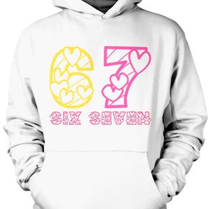 May include: White hoodie with a drawstring hood and a kangaroo pocket. The front features the number "67" in yellow and pink, with heart-shaped cutouts. The text "SIX SEVEN" is printed below in pink.