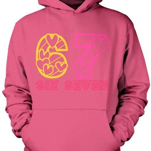 May include: Pink hooded sweatshirt with the number 67 in yellow and pink, with heart-shaped designs. The words "SIX SEVEN" are printed below the numbers. The sweatshirt has a front pocket and a hood.