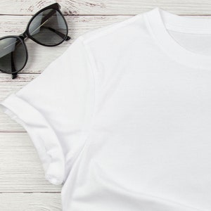 Puede incluir: Camiseta blanca con mangas enrolladas sobre un fondo de madera blanca. Un par de gafas de sol negras se encuentra en la esquina superior izquierda de la imagen.