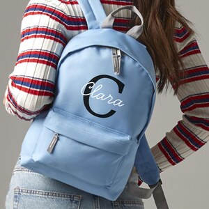 Può includere: Uno zaino azzurro con una tasca frontale e una maniglia superiore. Lo zaino presenta il nome "Clara" in corsivo bianco all'interno di una "C" nera sul davanti. Chiusura con zip e spallacci regolabili.