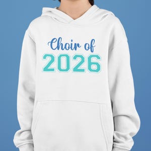 Op de afbeelding: Witte hoodie met de tekst "Choir of 2026" in blauw en teal. De sweatshirt heeft een voorzak en een capuchon met trekkoord. De achtergrond is effen blauw.
