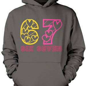 May include: Dark gray hoodie with a hood and a front pocket. The front features the number "67" in yellow and pink, with heart-shaped cutouts. Below the numbers, the words "SIX SEVEN" are printed in pink.