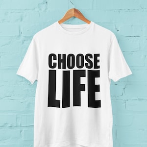 May include: White t-shirt with black text that reads "CHOOSE LIFE" hanging on a wooden hanger against a light blue brick wall.