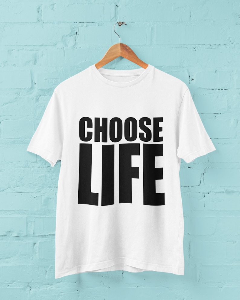 May include: White t-shirt with black text that reads "CHOOSE LIFE" hanging on a wooden hanger against a light blue brick wall.