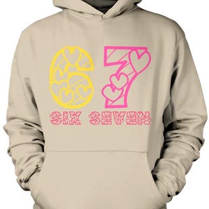 May include: A beige hoodie with a hood and a front pocket. The design features the number "67" in yellow and pink, with heart-shaped details. The words "SIX SEVEN" are printed below the numbers in pink.