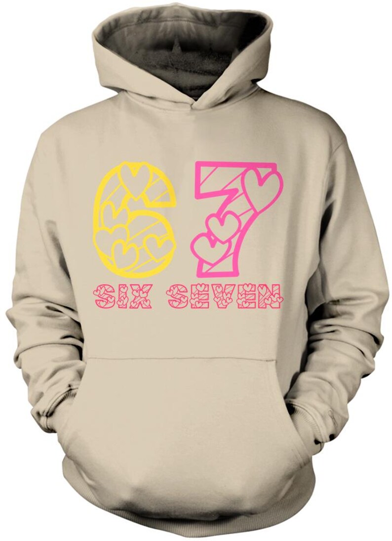 May include: A beige hoodie with a hood and a front pocket. The design features the number "67" in yellow and pink, with heart-shaped details. The words "SIX SEVEN" are printed below the numbers in pink.