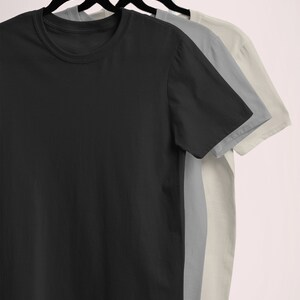 Puede incluir: Tres camisetas colgadas en un perchero blanco. Las camisetas son negra, gris y beige.