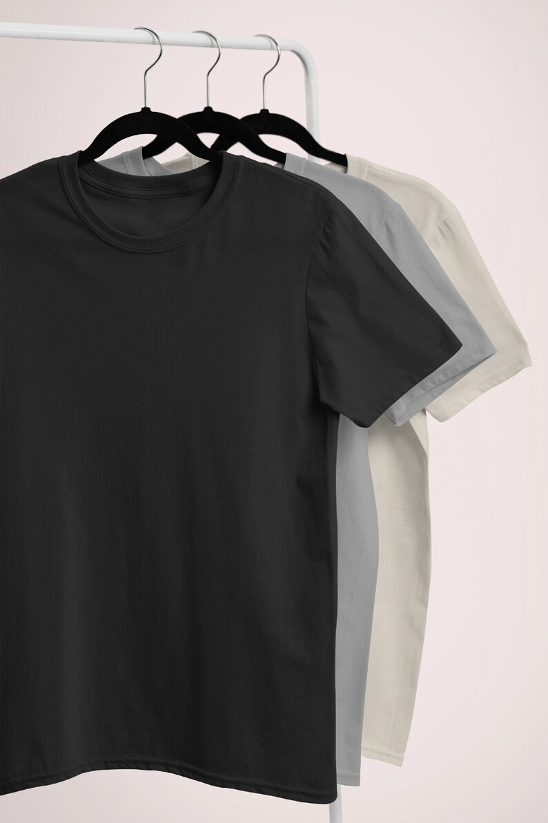 Puede incluir: Tres camisetas colgadas en un perchero blanco. Las camisetas son negra, gris y beige.