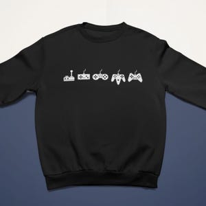 Peut inclure: Sweat-shirt noir avec un motif blanc représentant l'évolution des manettes de jeux vidéo. Le design montre la progression d'un joystick à une manette moderne. Un vêtement confortable et élégant pour les joueurs.
