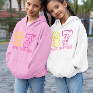 May include: Two children wearing pink and white hoodies with the number "67" in yellow and pink, and the words "SIX SEVEN" below. The hoodies have a drawstring hood and a front pocket. The children are wearing blue jeans.