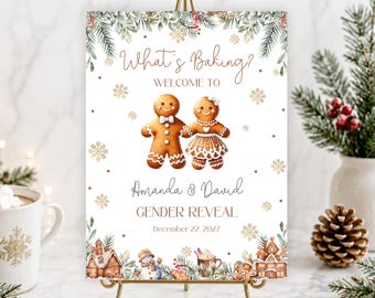 May include: A rectangular sign on a gold metal stand featuring the text What's Baking? Welcome to Amanda & David Gender Reveal December 22 2027. The design includes two gingerbread cookies, pine branches, snowflakes, and gingerbread houses.