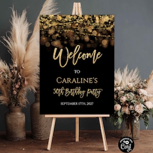 May include: A rectangular welcome sign on a wooden easel featuring a black background with gold bokeh lights at the top. The text reads: Welcome TO CARALINE'S 50th Birthday Party SEPTEMBER 17TH, 2027. The sign has gold script and serif lettering.