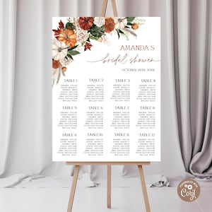 May include: A white bridal shower seating chart on an easel, featuring floral accents of pumpkins and foliage. The chart displays guest names organized by table numbers, with the text "Amanda's bridal shower" at the top.