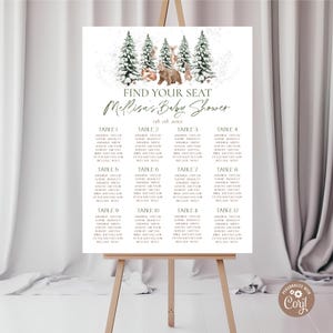 May include: A white baby shower seating chart on an easel. The top features a watercolor illustration of pine trees and woodland animals. The text reads "Find Your Seat" and "Melissa's Baby Shower" with the date. Twelve tables are listed.