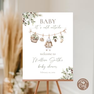 May include: A baby shower welcome sign with the text "BABY it's cold outside." The sign features watercolor illustrations of winter-themed decorations, including a basket, a coat, and a stocking. The sign is on a copper-coloured easel.