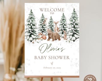 May include: A welcome sign for a baby shower, featuring a winter forest scene with snow-covered pine trees and woodland animals. The sign reads "Welcome to Olivia's Baby Shower" with the date February 20, 2025.