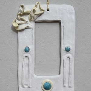May include: White ceramic picture frame with a rectangular aperture, decorated with a cream-coloured floral accent and three turquoise-coloured circular embellishments. The frame is suspended by natural twine.