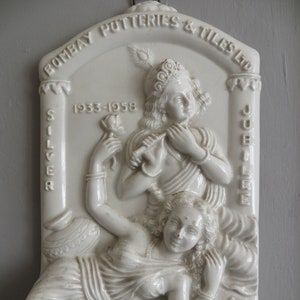 Puede incluir: Placa de cerámica blanca con una escultura en relieve de una pareja. La placa está decorada con el texto "Bombay Potteries & Tiles Ltd. Silver Jubilee 1933-1958".