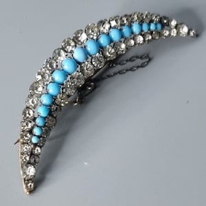 May include: A crescent-shaped brooch with a silver tone finish. The brooch is adorned with a row of turquoise blue stones and a row of clear rhinestones.