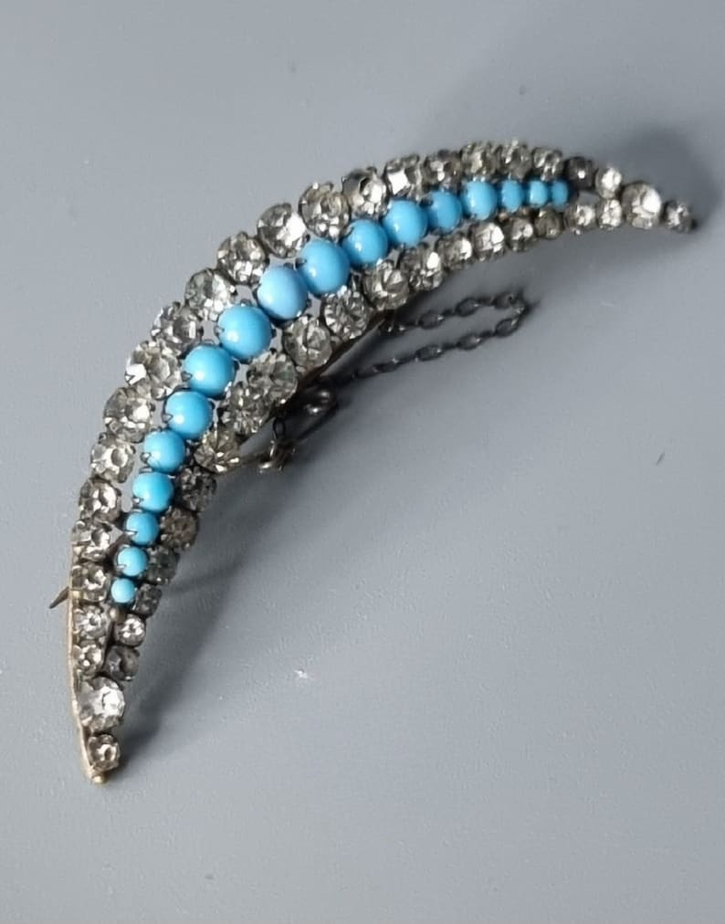 May include: A crescent-shaped brooch with a silver tone finish. The brooch is adorned with a row of turquoise blue stones and a row of clear rhinestones.