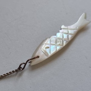 Puede incluir: Un amuleto de pez de nácar blanco con un diseño tallado detallado. El amuleto está unido a una cadena dorada.